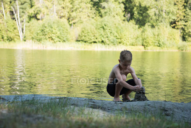 Menino brincando com areia perto da margem do rio em um dia ensolarado — Fotografia de Stock