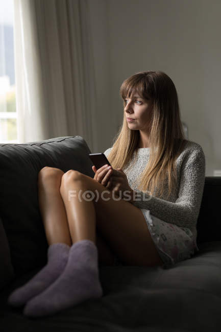Thoughtful woman holding mobile phone in living room at home. — Stock Photo