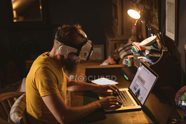 Homem sentado na cadeira vestindo fone de ouvido realidade virtual usando seu laptop em casa — Fotografia de Stock