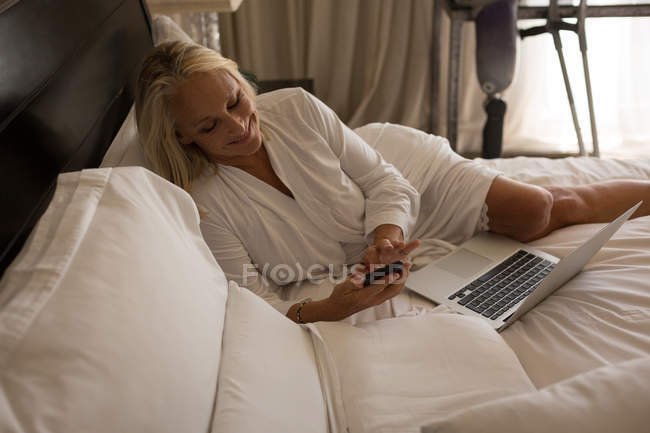 Жінка з обмеженими можливостями, використовуючи мобільний телефон і ноутбук на ліжку вдома . — стокове фото
