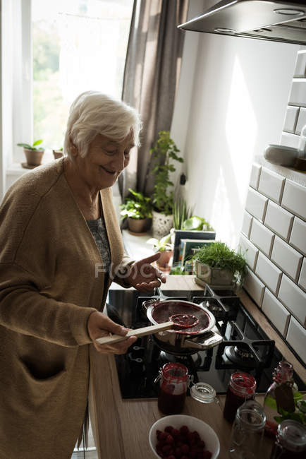 Sorrindo mulher idosa cozinhar geléia de framboesa na cozinha em casa — Fotografia de Stock