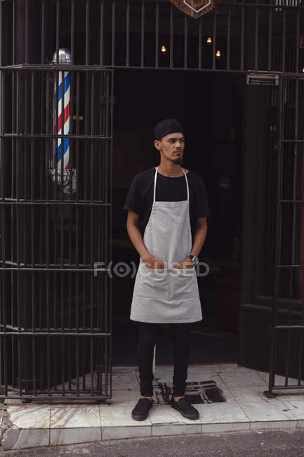 Задумчивый парикмахер стоит с руками в кармане у входа в свой магазин — стоковое фото