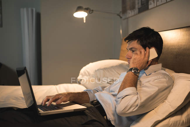 Empresario que utiliza el ordenador portátil mientras habla por teléfono móvil en el dormitorio - foto de stock