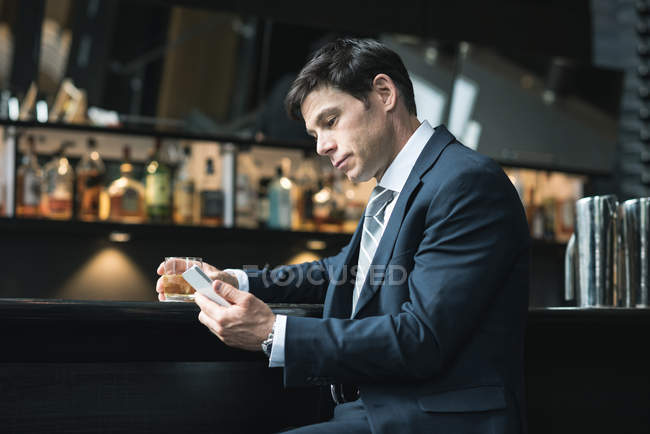 Empresário usando telefone celular enquanto bebe uísque no balcão do hotel — Fotografia de Stock