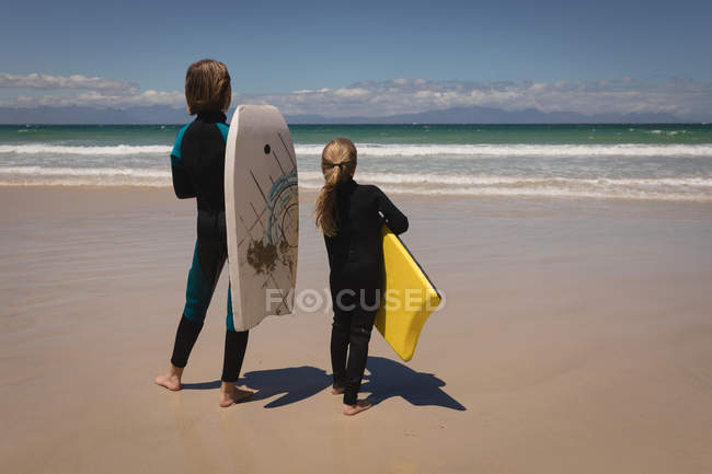 Vista traseira dos irmãos de fato de mergulho em pé com prancha de surf na praia — Fotografia de Stock