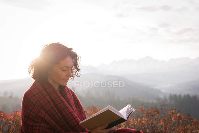 Mujer envuelta en libro de lectura de manta en día de niebla - foto de stock