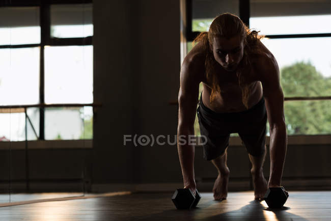 Young man with long red hair doing push-ups with dumbbells in fitness studio. — Stock Photo