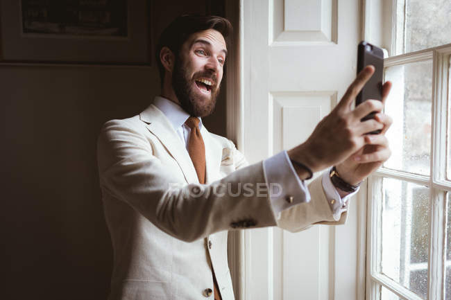 Emocionado novio tomando una selfie cerca de la ventana en casa - foto de stock