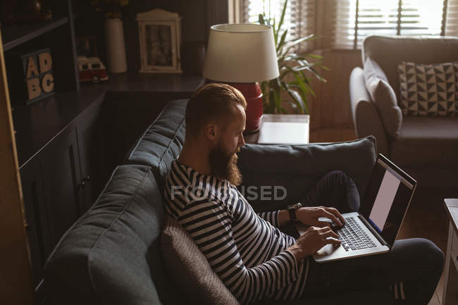 Vista lateral del hombre sentado en el sofá usando su computadora portátil - foto de stock