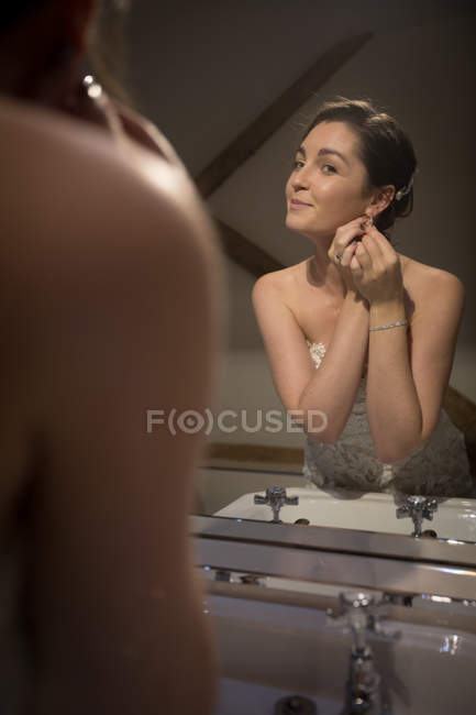 Улыбающаяся невеста носит серьги перед зеркалом дома — стоковое фото