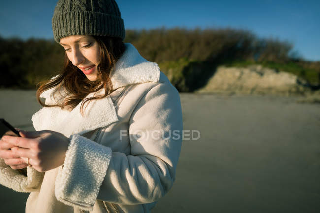 Jeune femme en bonnet de laine en utilisant le téléphone mobile sur le bord de la rivière . — Photo de stock