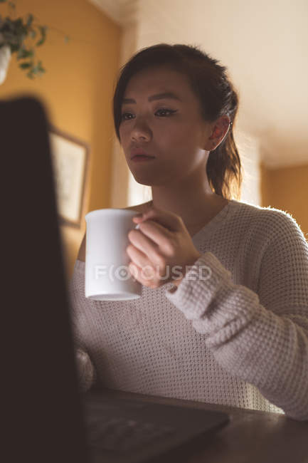 Femme utilisant un ordinateur portable tout en prenant un café à la maison — Photo de stock