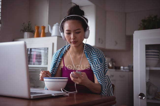 Femme écoute de la musique sur téléphone portable tout en prenant le petit déjeuner à la maison — Photo de stock