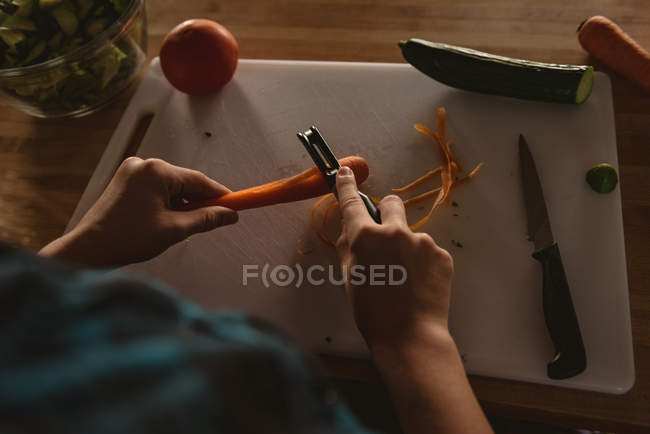 Vista ad alto angolo di ragazza in piedi in cucina e peeling carota con pelapatate . — Foto stock