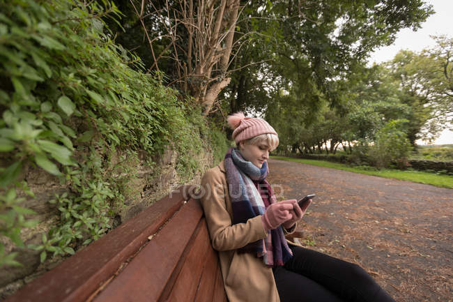 Jovem mulher sentada no banco usando seu telefone celular no parque — Fotografia de Stock