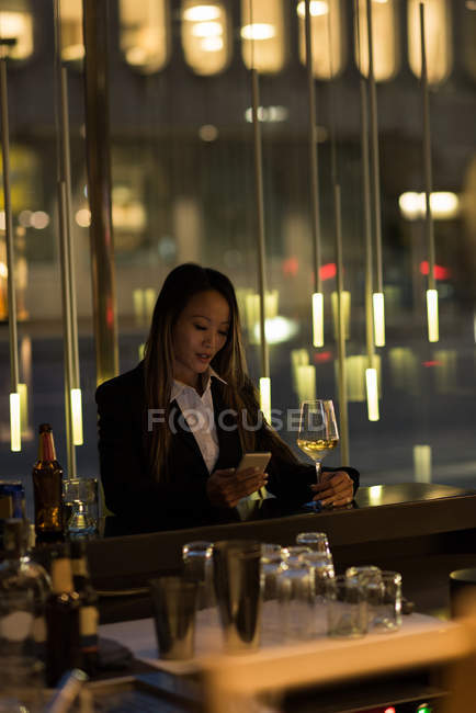 Businesswoman standing at bar having champagne while using her phone in the hotel — Stock Photo