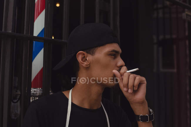 Jovem barbeiro fumando cigarro na entrada de sua loja — Fotografia de Stock