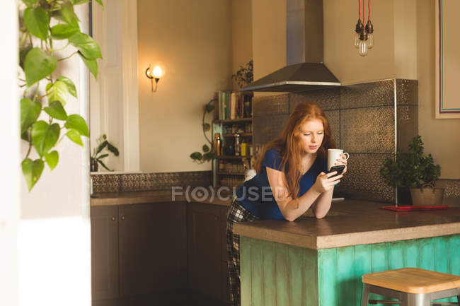 Femme prenant un café tout en utilisant un téléphone portable à la maison — Photo de stock