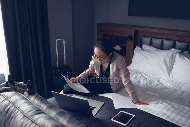 Mujer leyendo documento mientras trabaja en el ordenador portátil en el hotel - foto de stock