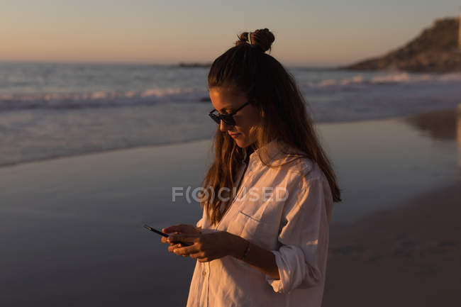 Молодая женщина использует мобильный телефон на пляже в сумерках . — стоковое фото