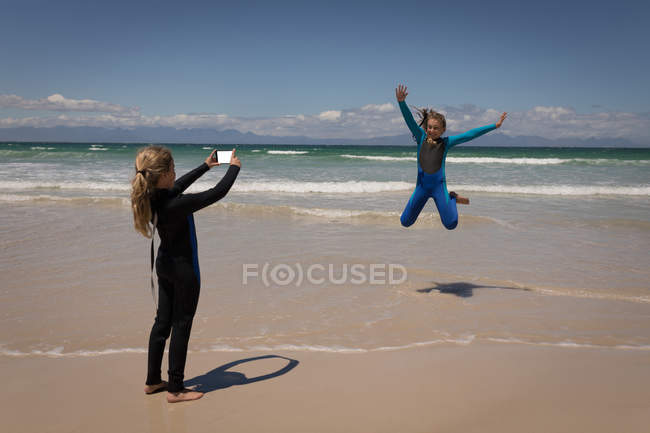 Girl taking picture of her sister with mobile phone on beach — Stock Photo