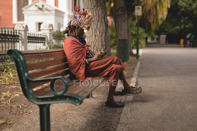 Maasai homem em roupas tradicionais usando telefone celular enquanto relaxa no banco — Fotografia de Stock