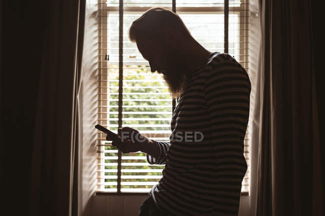 Чоловік стоїть біля вікна, використовуючи свій мобільний телефон в домашніх умовах — стокове фото