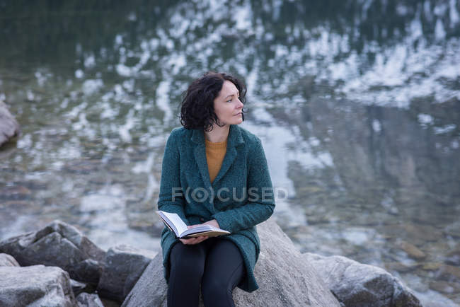 Mujer pensativa sentada con libro a orillas del lago durante el invierno - foto de stock