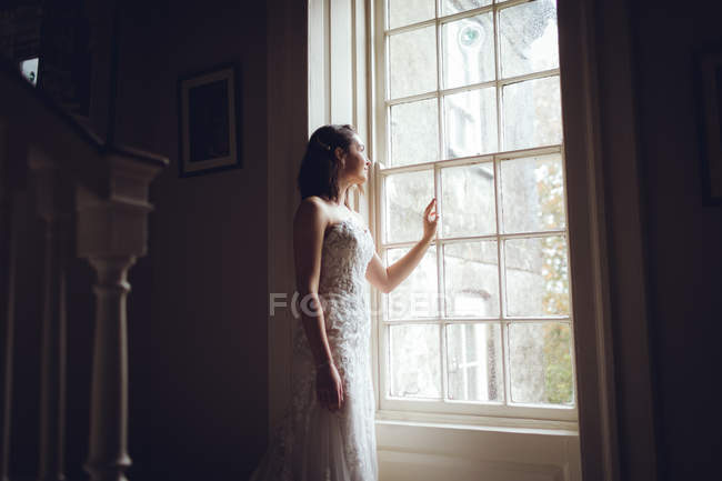 Dreamy bride looking out of the window at home — Stock Photo