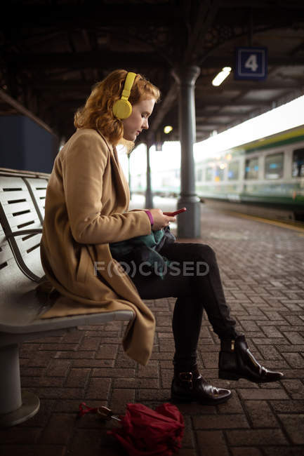 Jovem mulher listando música ao usar seu telefone celular na plataforma ferroviária — Fotografia de Stock