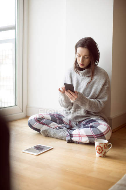 Молода жінка використовує мобільний телефон, сидячи з ногами, схрещеними на підлозі вдома . — стокове фото