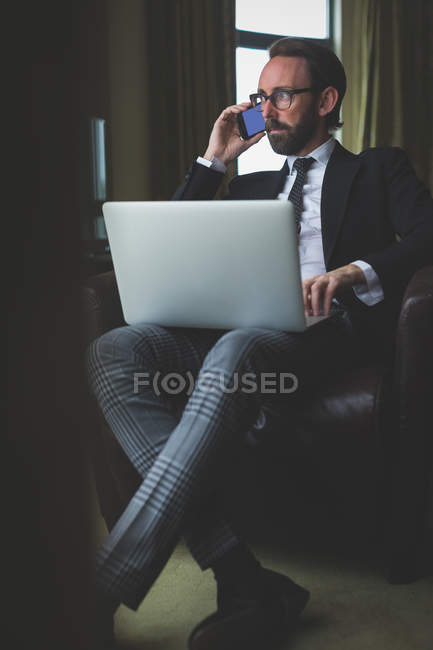 Homme d'affaires parlant sur un téléphone portable tout en utilisant un ordinateur portable dans la chambre d'hôtel — Photo de stock