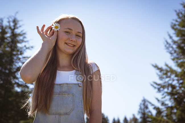 Menina sorridente colocando flor no cabelo no campo à luz do sol . — Fotografia de Stock