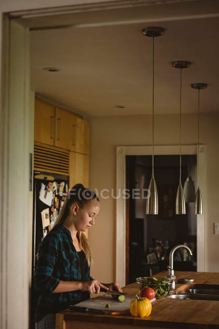 Menina de pé na cozinha e corte de legumes com faca em casa . — Fotografia de Stock