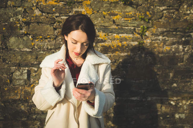 Woman using mobile phone against stone wall in sunlight. — Stock Photo