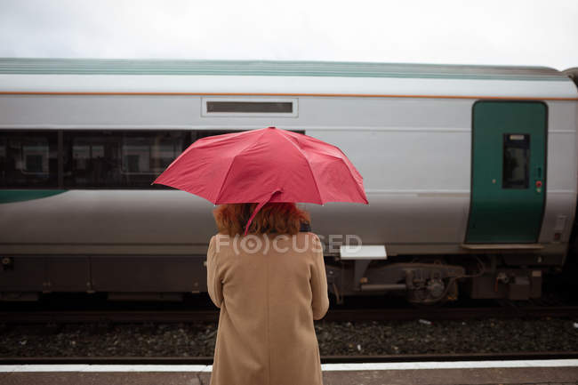 Vue arrière de belle jeune femme avec parapluie attendant le train sur quai — Photo de stock