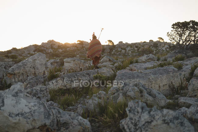 Vista trasera del hombre maasai con ropa tradicional caminando sobre roca en el campo - foto de stock