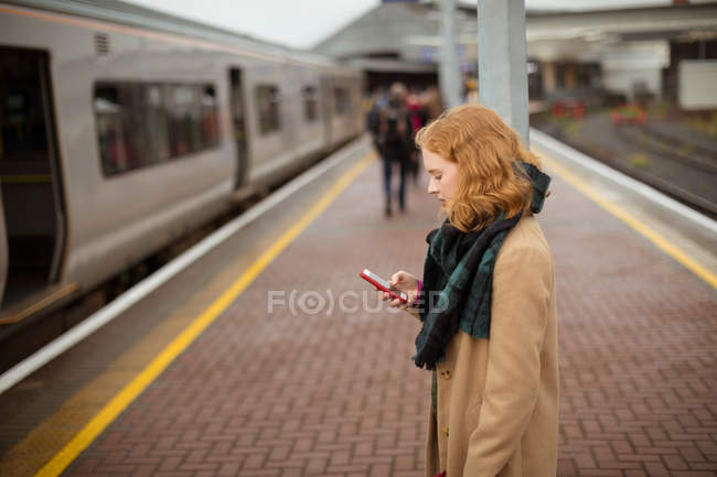 Jovem mulher de pé na plataforma ferroviária usando seu telefone celular em um dia chuvoso — Fotografia de Stock