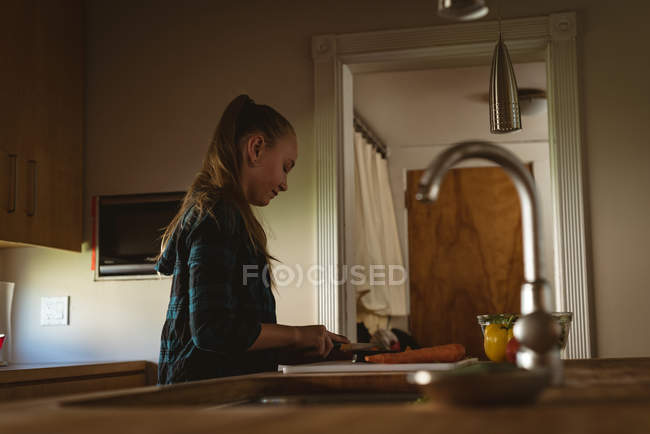 Vista lateral de la niña de pie en la cocina y el corte de verduras con cuchillo en casa . - foto de stock