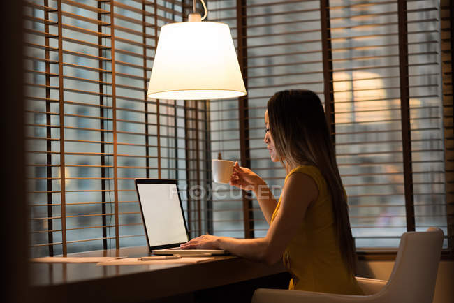 Empresária tomando café enquanto trabalhava em laptop no escritório — Fotografia de Stock