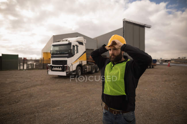 Thoughtful dock worker adjusting his hard hat in the shipyard — Stock Photo