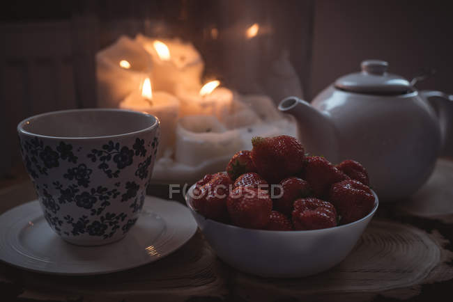 Primer plano de la tetera, taza y tazón de fresas contra velas encendidas - foto de stock