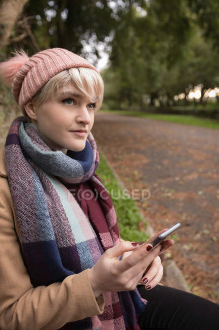 Продумана жінка в теплому одязі, використовуючи мобільний телефон у парку — стокове фото