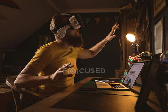 Hombre con auriculares de realidad virtual sentado frente a la computadora portátil en casa - foto de stock