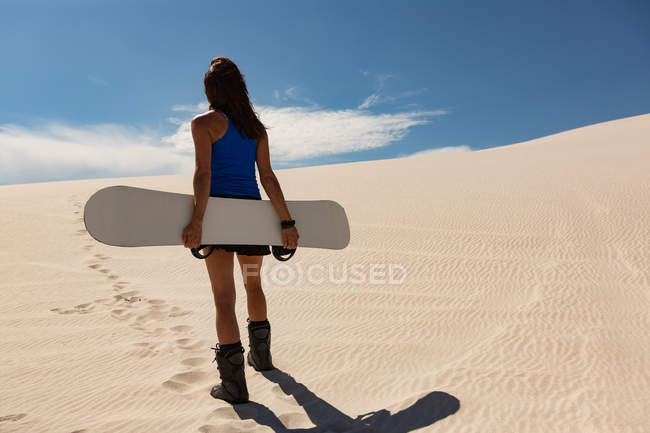 Вид сзади женщины с песчаной доской, стоящей в пустыне в солнечный день — стоковое фото