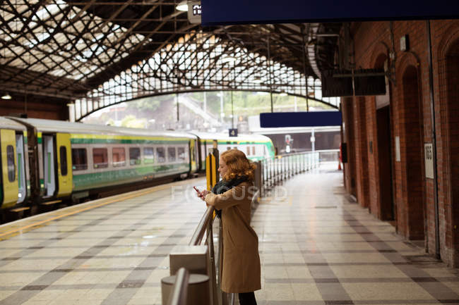Молода жінка стоїть на залізничній платформі, використовуючи свій мобільний телефон — стокове фото
