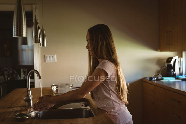 Menina de pé na cozinha e lavar as mãos sob água da torneira em casa . — Fotografia de Stock