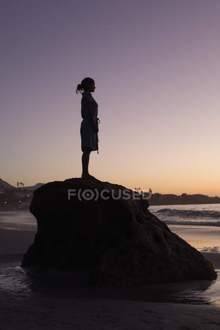 Silhouette di donna in piedi su roccia in spiaggia al tramonto . — Foto stock