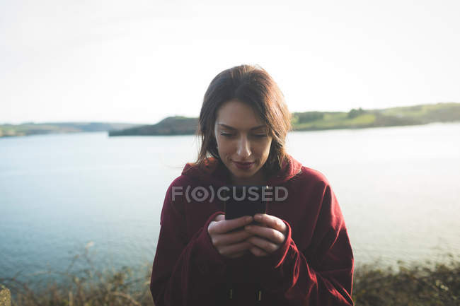 Mujer usando teléfono móvil cerca de la orilla del río a la luz del sol . - foto de stock