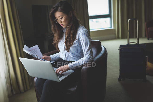 Empresária segurando documentos enquanto trabalhava em laptop no quarto de hotel — Fotografia de Stock